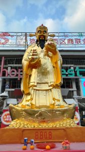 A grand golden statue of the Chinese God of Wealth, Caishen, standing majestically with gold ingots and a prosperity urn, surrounded by festive decorations and offerings at a marketplace.
