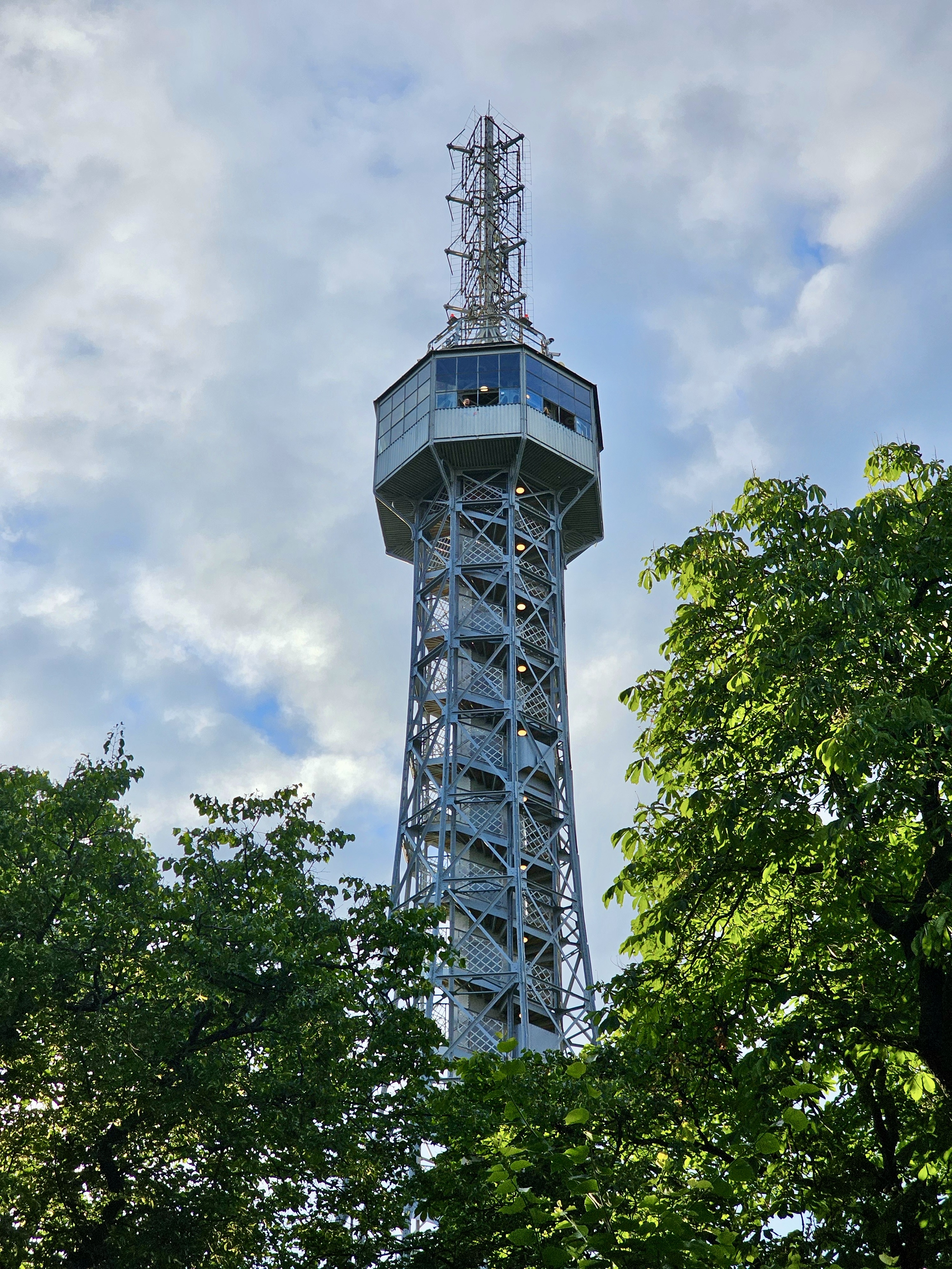 The Petřín Lookout Tower in Prague, Czech Republic, surrounded by green trees, with a cloudy sky in the background.