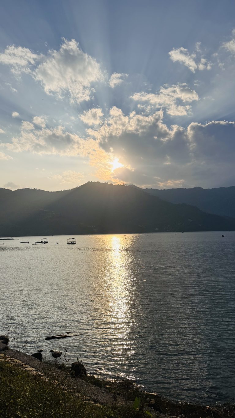 Sunset over a fewa lake pokhara with boats scattered on the water. The sun is partially obscured by clouds, casting rays and a reflection on the water, surrounded by silhouetted hills. In the foreground, there’s a grassy shoreline.