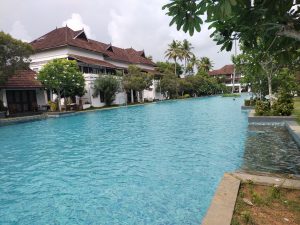 A large, clear swimming pool extends along a row of white buildings with brown tiled roofs, surrounded by lush green trees and plants. The sky is overcast, with some clouds visible.