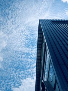 A low-angle view of the corner of a modern building with reflective windows, set against a sky filled with scattered, fluffy clouds.
