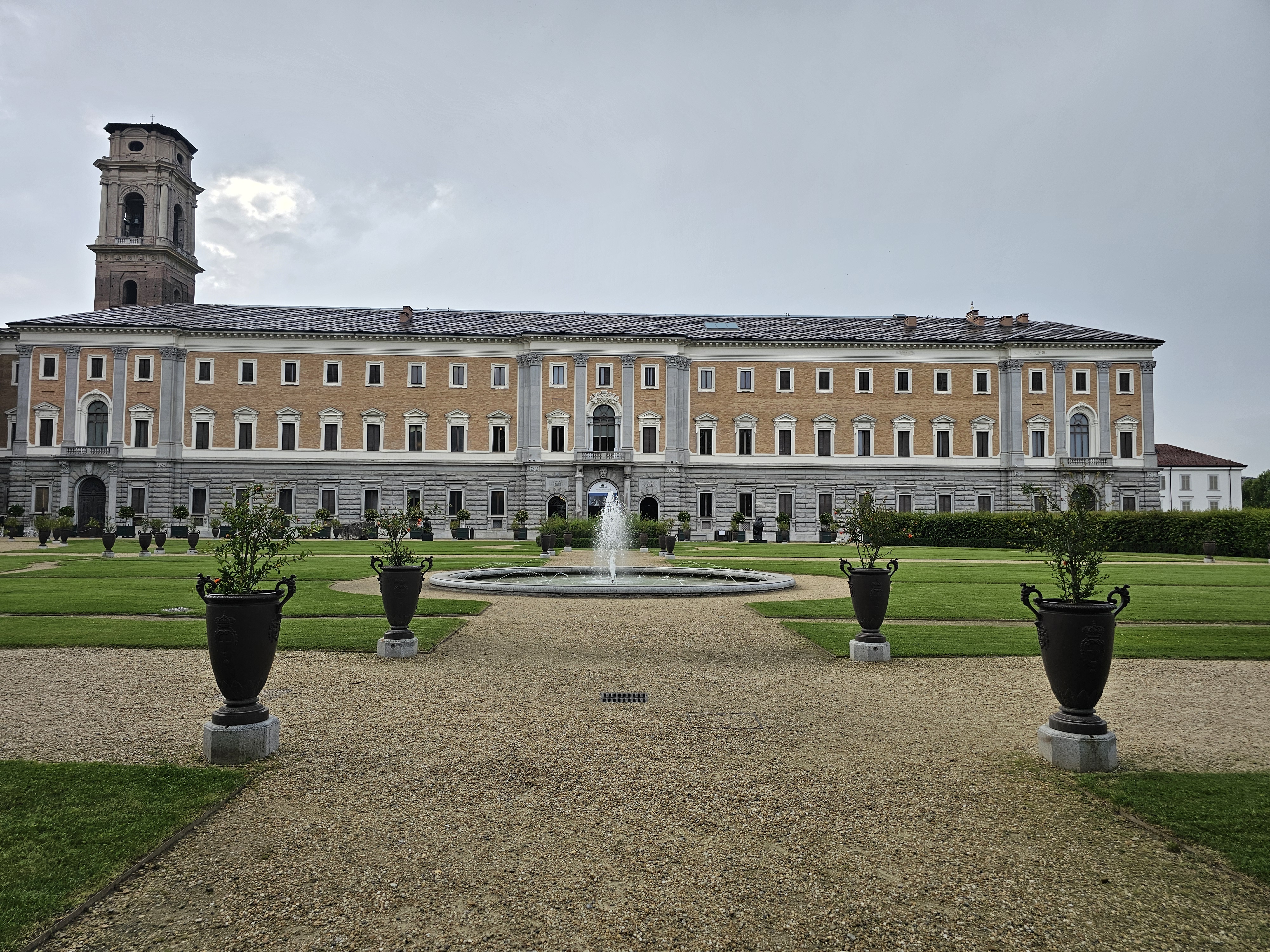 The Royal Palace of Turin (Palazzo Reale) with its gardens (Giardini Reali) and the Galleria Sabauda. The palace is a large, symmetrical building with a tower on the left side. It has a pale yellow facade with rows of windows and a dark roof.