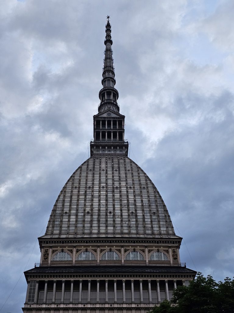 A long view of the upper part of the Mole Antonelliana in the evening, a major landmark in Turin, Italy, featuring its distinctive dome and spire against a cloudy sky. The architectural details of the building are prominently displayed, emphasizing its unique design.