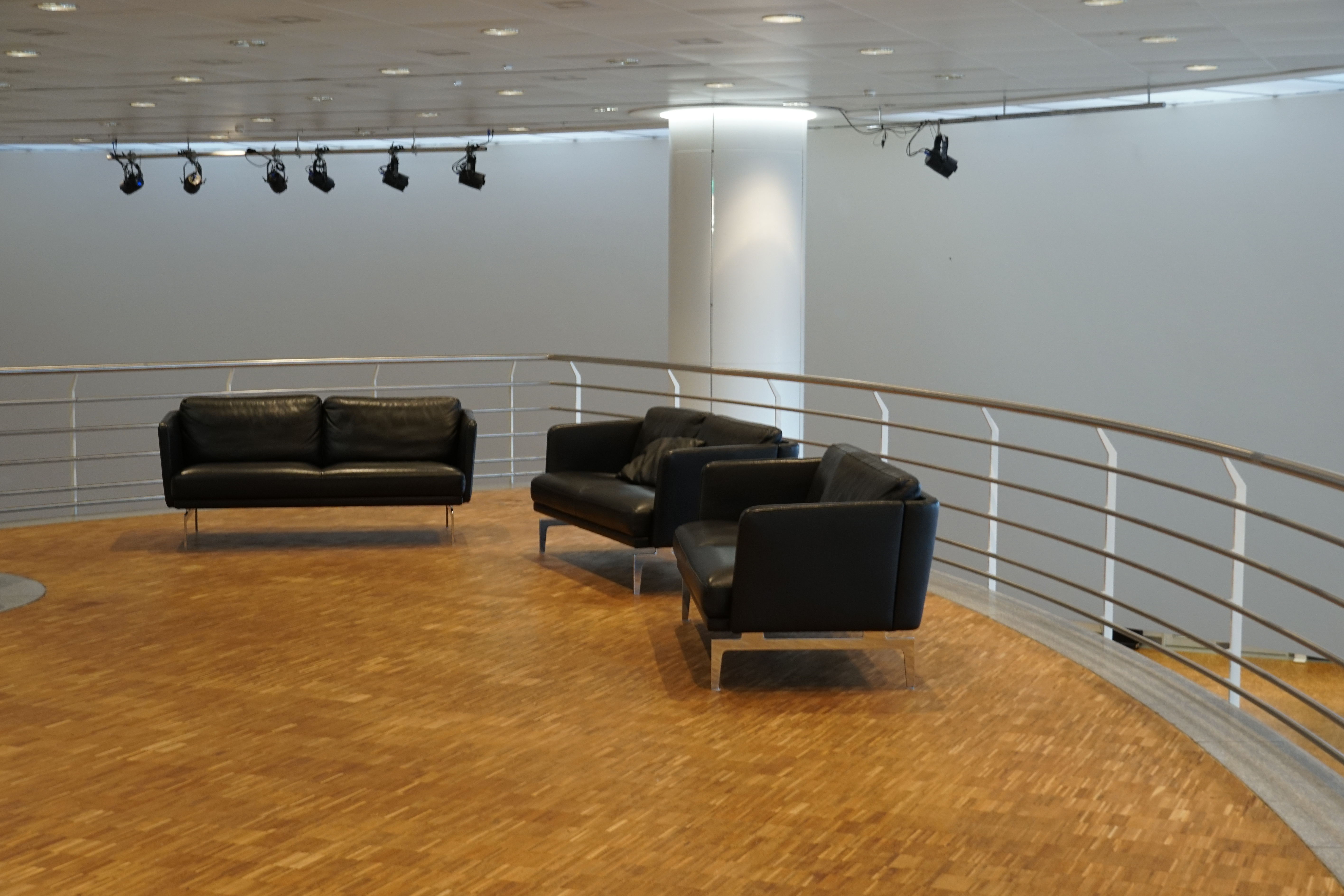 A modern indoor waiting area with wooden flooring, featuring two black leather sofas and a black armchair. The space is illuminated with overhead spotlights and bordered by a silver metal railing.