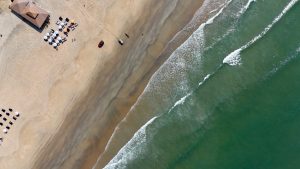 Aerial view of Cavelossim Beach in South Goa, showing a stretch of golden sand meeting the greenish-blue ocean waves. Gentle waves create a foamy white pattern as they roll onto the shore. A cluster of beach umbrellas and lounge chairs is arranged near a large shaded structure. Tire tracks and footprints are visible in the sand.
