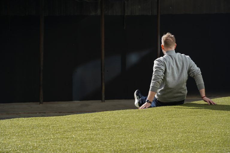 A person wearing a gray sweater and jeans is sitting on a grassy slope, facing away from the camera. The background is dark and shadowed, and the person appears to be relaxed with one hand on the ground for support.