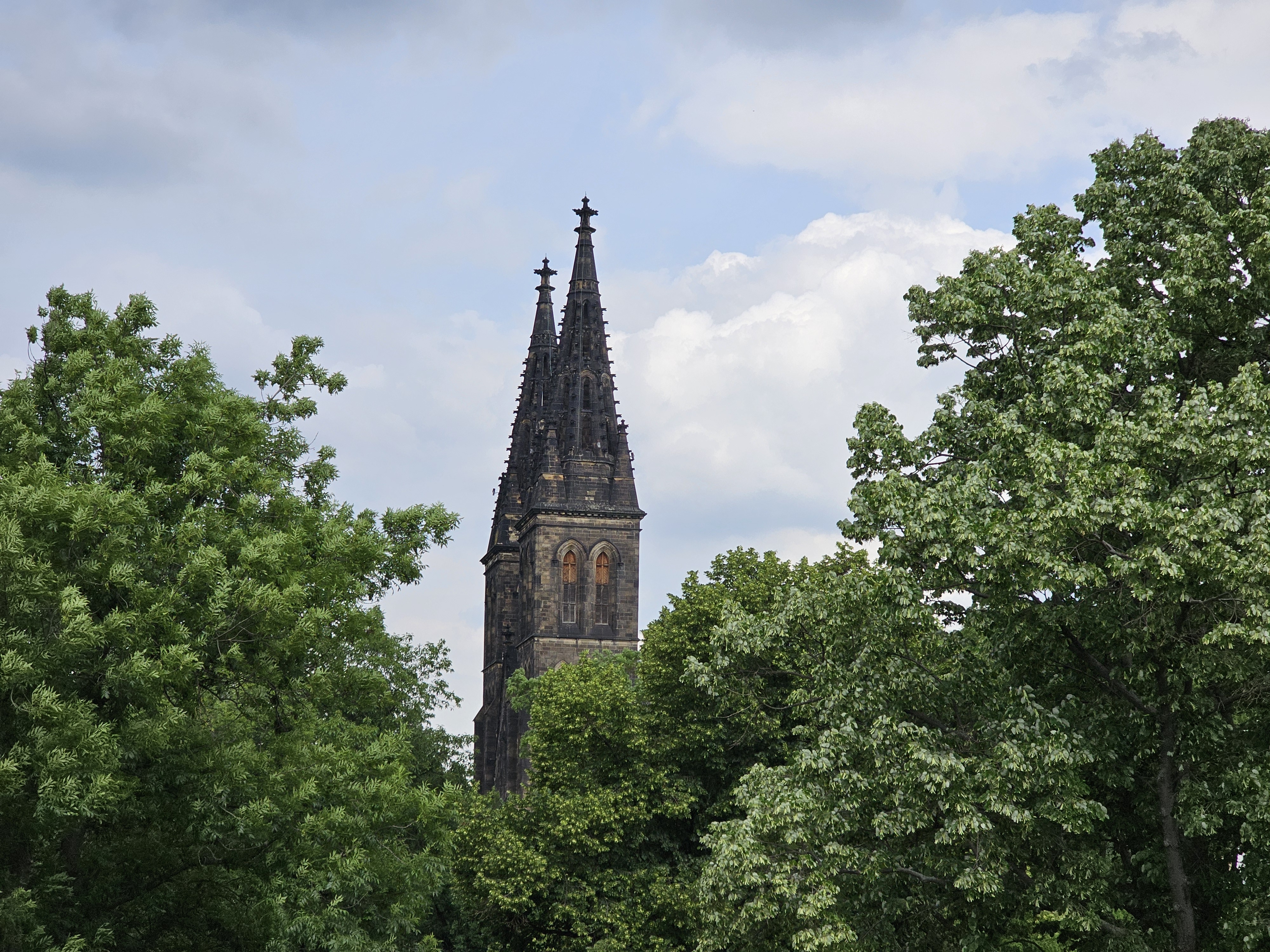 The Basilica of St. Peter and St. Paul in Vyšehrad, Prague, with its two prominent spires rising above the surrounding trees. The basilica's dark stone contrasts with the green foliage, and the sky is partly cloudy.