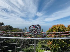 A heart-shaped metal structure adorned with colorful padlocks on a scenic viewpoint railing, overlooking lush green trees and an ocean horizon under a bright blue sky with scattered clouds.