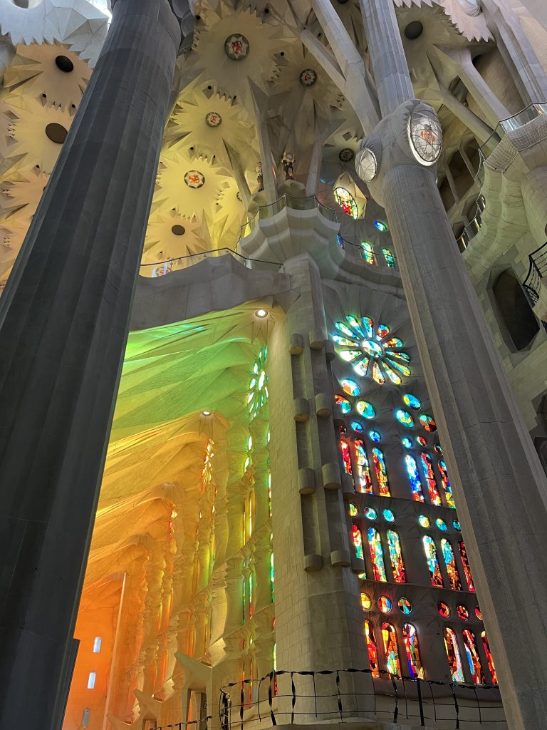 The interior of of the Sagrada Familia cathedral with tall pillars and intricate ceiling designs. Stained glass windows cast colorful light patterns in shades of red, blue, green, and yellow throughout the space.