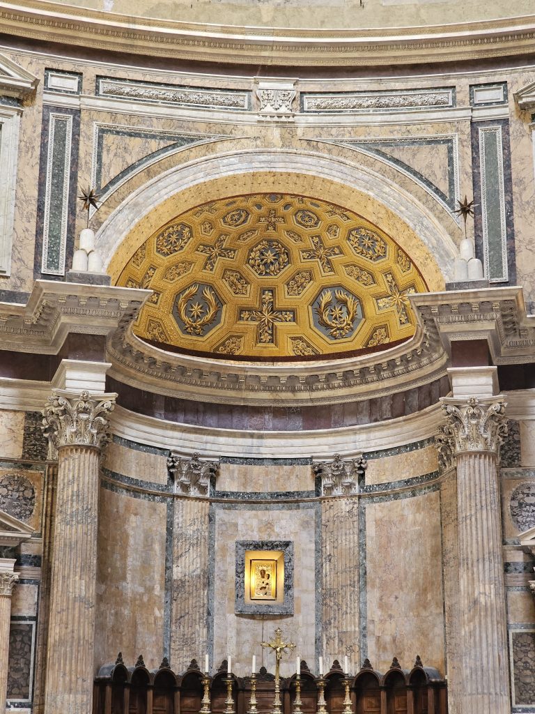 The interior of the Pantheon in Rome, Italy. The view is focused on a semi-domed apse area featuring a golden, coffered ceiling with geometric patterns and crosses. Below the apse, there is a framed artwork, likely a religious icon, positioned centrally on the wall.