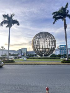 A large spherical sculpture stands in the center of the image, surrounded by grass and positioned near a modern building.

