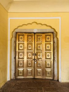 Ornate golden wooden doors with decorative metal inlays, set within a yellow arched doorway framed by a scalloped border and white trim.