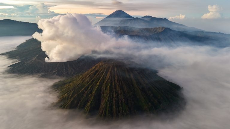 Aerial view of a volcanic landscape with a smoking crater surrounded by layers of mist and clouds. Several other volcanoes and mountain peaks are visible in the background, creating a dramatic scene under a clear sky.