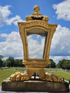 A large, ornate golden lantern with a metal glass pane stands on a stone base. It is set outdoors against a backdrop of a manicured green lawn, trees, and a partly cloudy blue sky. The lantern's design features decorative elements and is part of the Nymphenburg Palace grounds in Munich, Germany.
