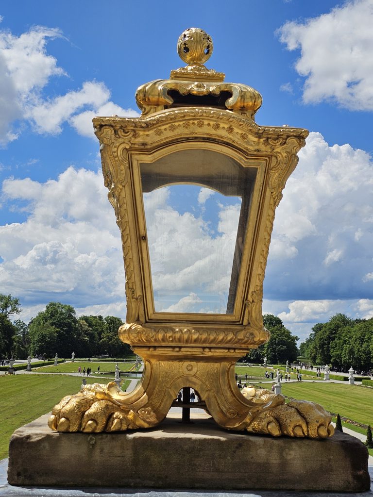 A large, ornate golden lantern with a metal glass pane stands on a stone base. It is set outdoors against a backdrop of a manicured green lawn, trees, and a partly cloudy blue sky. The lantern’s design features decorative elements and is part of the Nymphenburg Palace grounds in Munich, Germany.