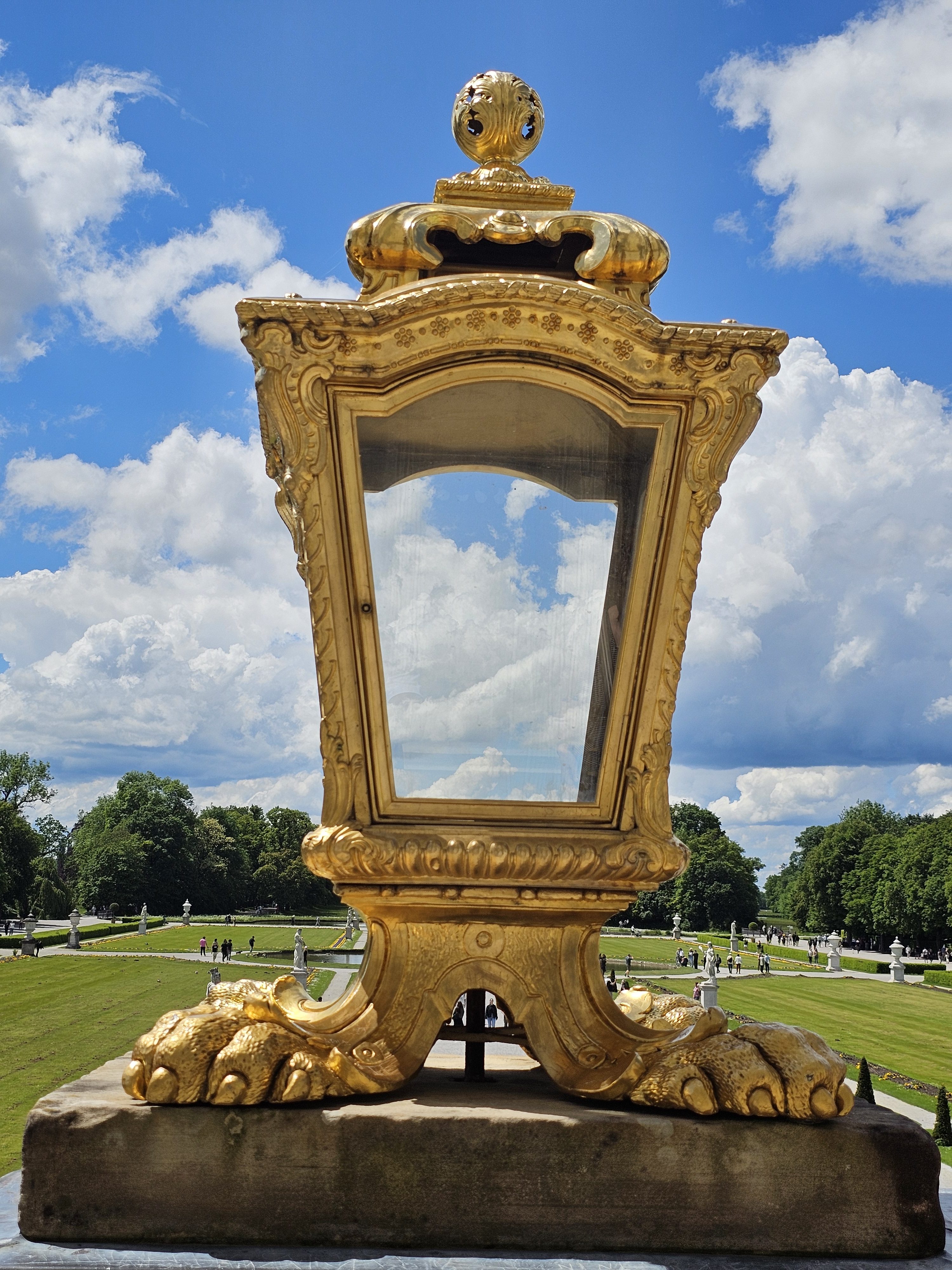 A large, ornate golden lantern with a metal glass pane stands on a stone base. It is set outdoors against a backdrop of a manicured green lawn, trees, and a partly cloudy blue sky. The lantern's design features decorative elements and is part of the Nymphenburg Palace grounds in Munich, Germany.