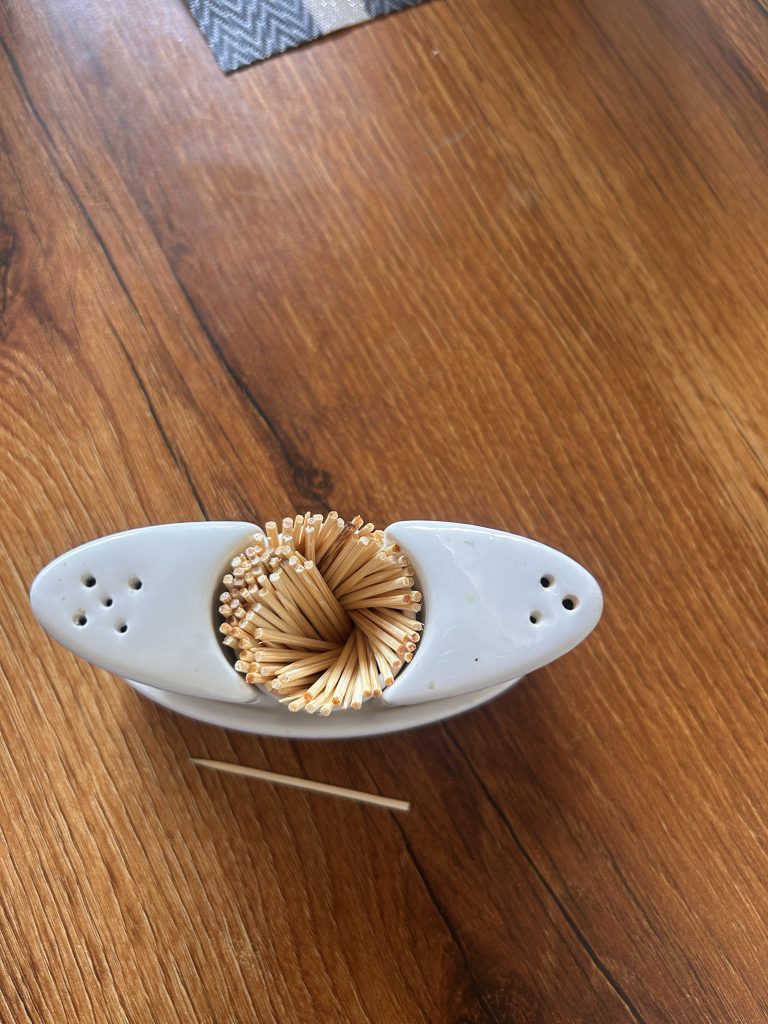 A white ceramic container holding a cluster of toothpicks on a wooden surface, with one toothpick placed beside it.