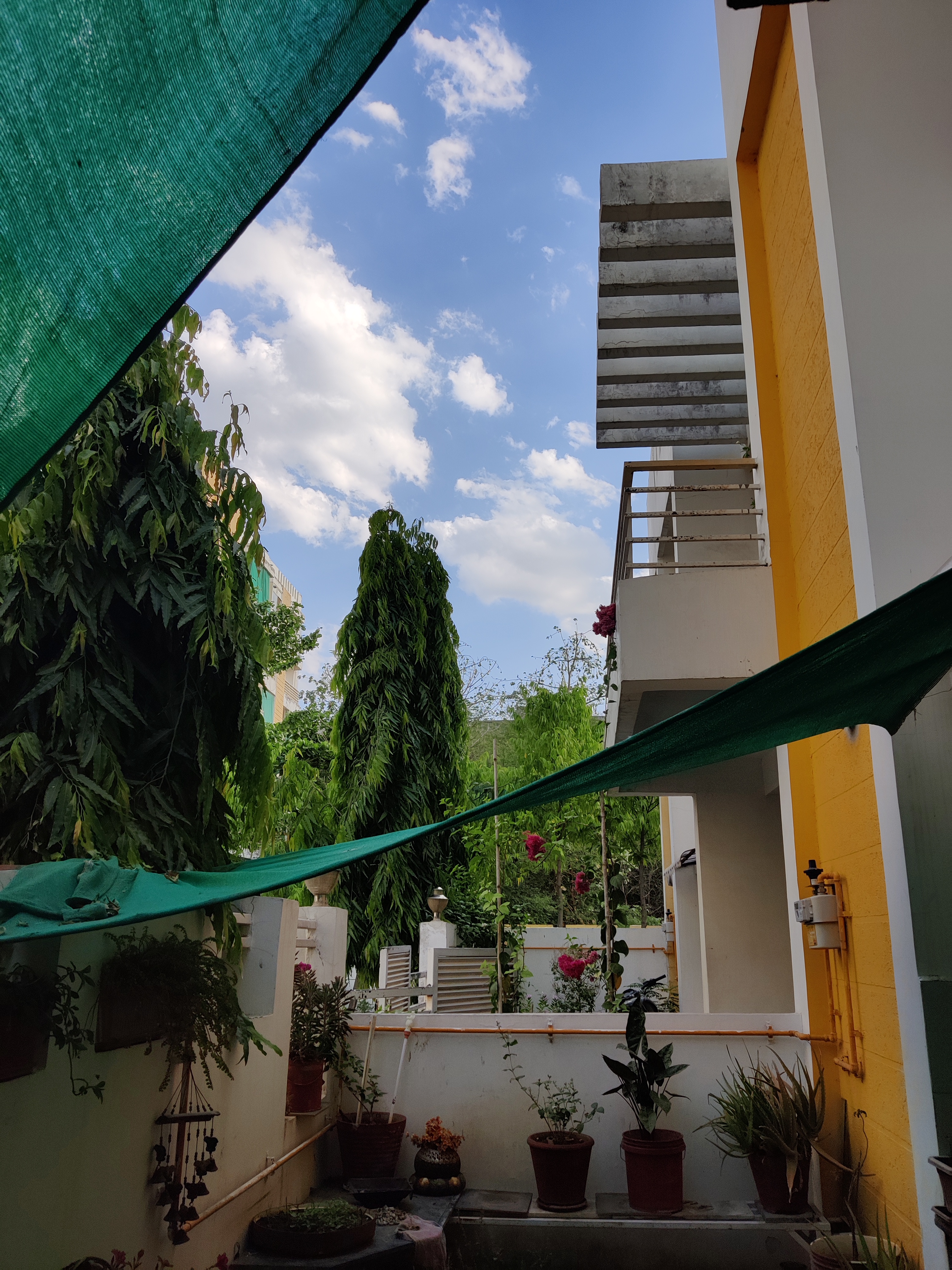 A view of a small outdoor area between buildings, featuring potted plants and greenery. A green shade cloth is stretched overhead.