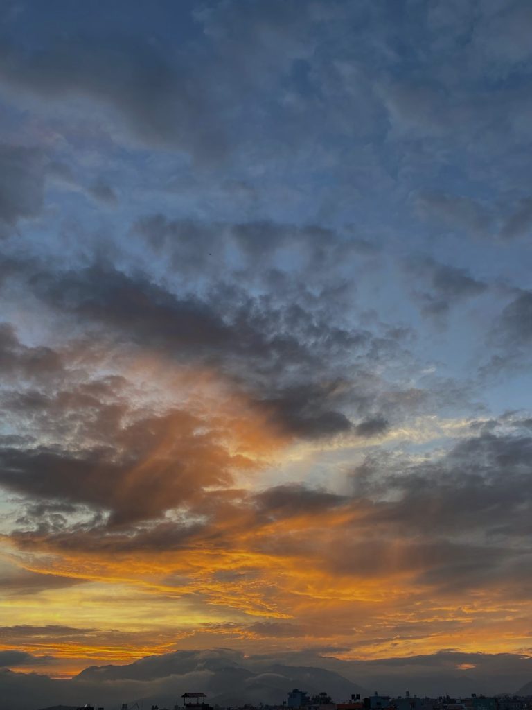 A vibrant sunset with a dramatic sky featuring dark clouds and a range of colors from deep orange to soft blue. The silhouette of a cityscape is visible at the bottom, with mountains in the distance.