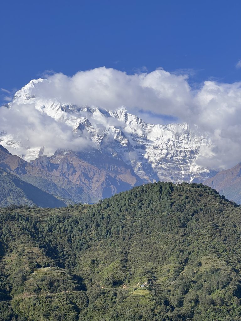 A lush green hillside is in the foreground, with a snow-capped mountain partially obscured by clouds in the background against a clear blue sky.