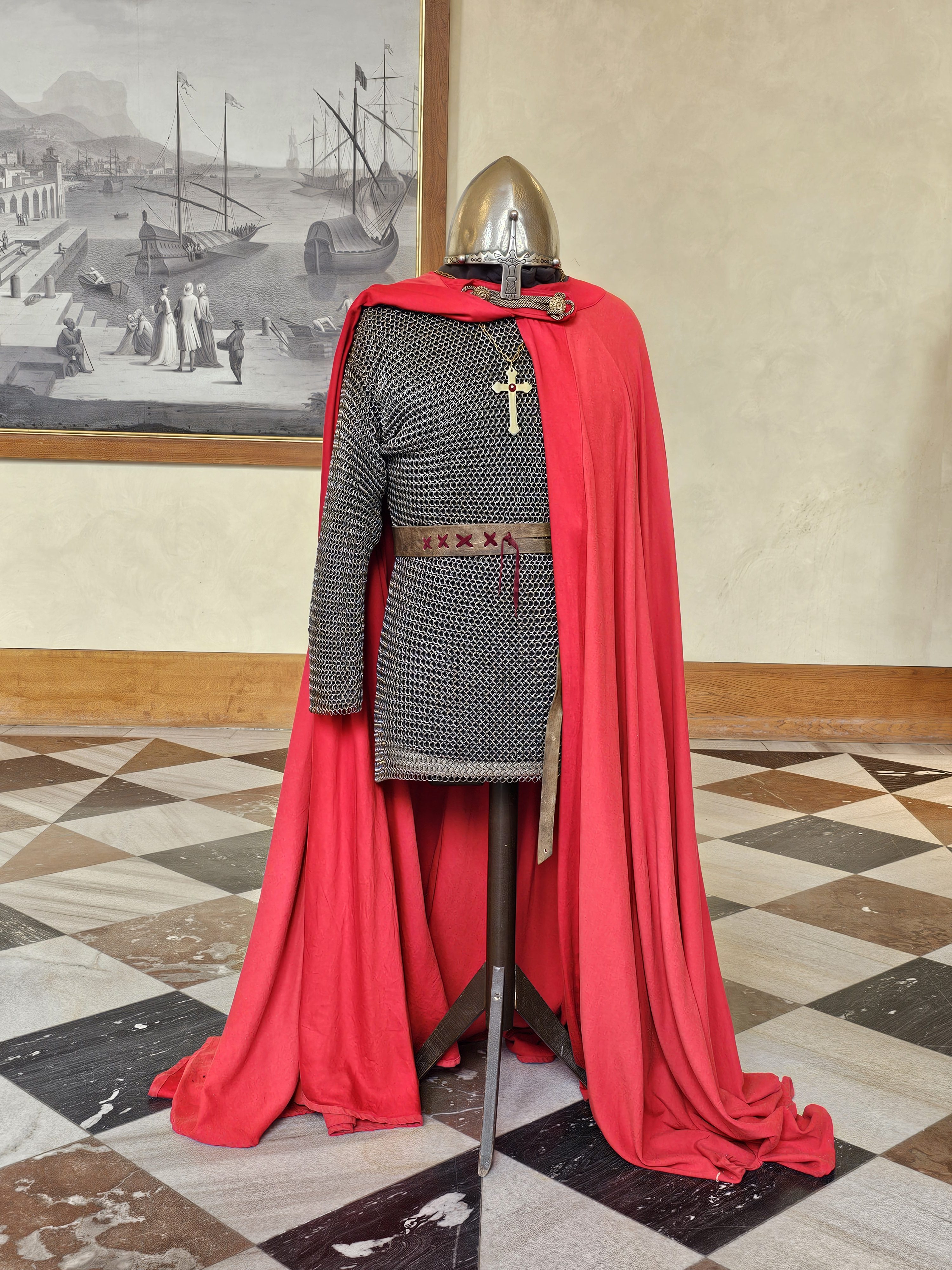 A medieval knight's attire, including a chainmail shirt, a red cape, and a helmet, is displayed on a stand. The outfit is complete with a belt and a cross emblem on the chest, set against a backdrop of a painting and a patterned floor. It was spotted from Prague Castle.