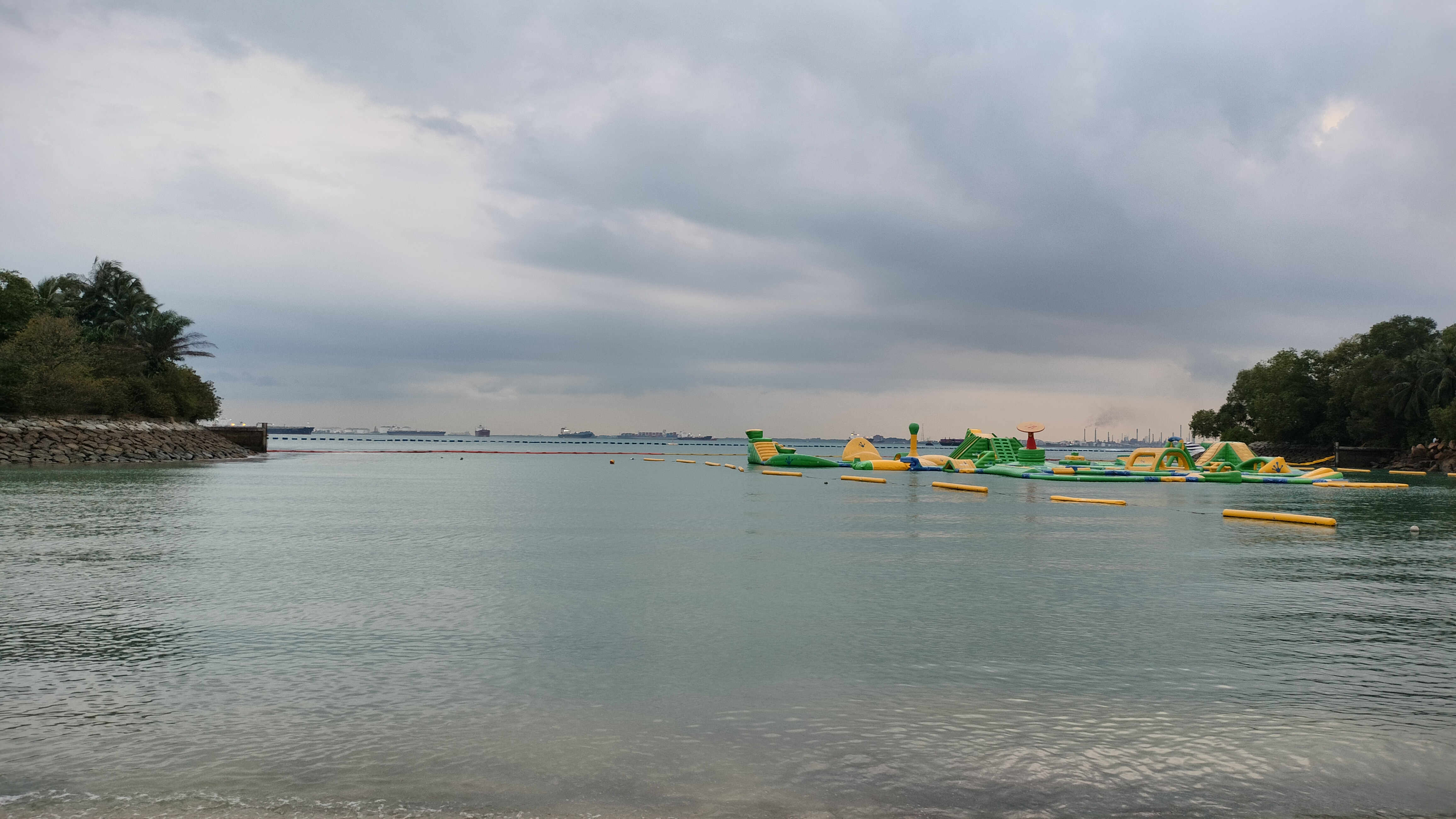 A calm body of water with an inflatable obstacle course floating on it. The backdrop features a cloudy sky, distant ships, and shoreline vegetation on either side.