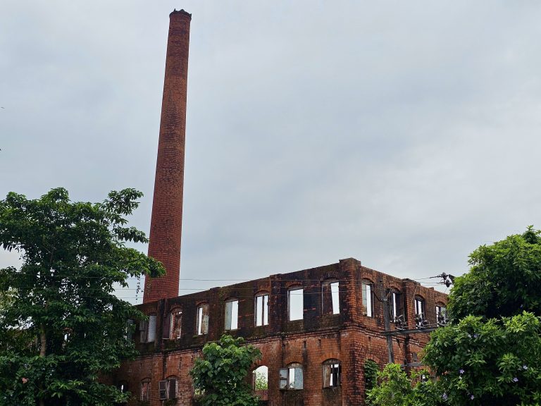 Long view of a ruined title factory. Its features a tall red brick chimney and a multi-story brick building with empty window frames. From Feroke, Kozhikode.