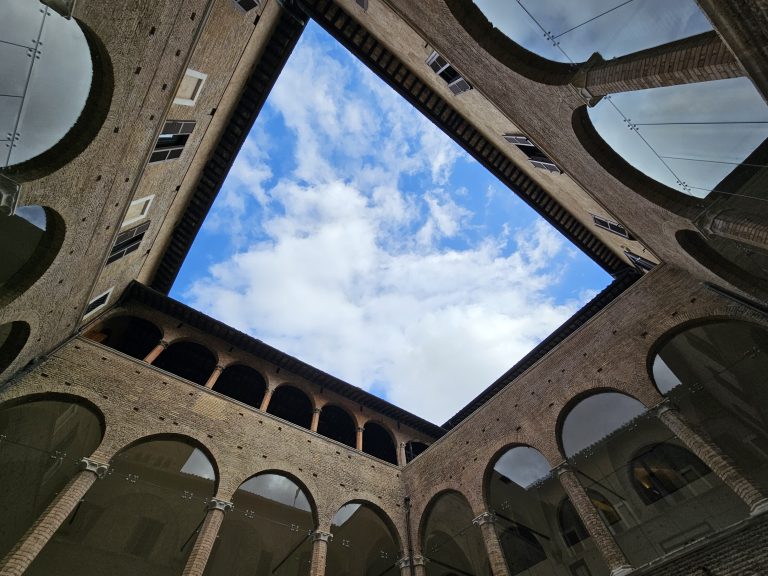 The internal courtyard of Santa Maria Antiqua in Rome, Italy, looking up at the sky. The courtyard is surrounded by brick walls with arched openings and columns, and the sky is visible in a square shape at the top of the frame. The sky is blue with some white clouds.