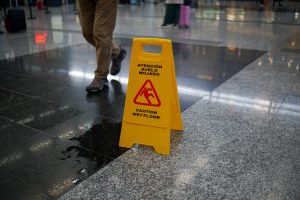 A yellow caution sign on a polished, wet floor in a public space, with the text "Caution Wet Floor" and its Spanish translation. A person walks nearby, partially visible in the frame.