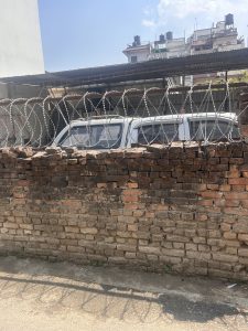 A brick wall topped with barbed wire, partially obscuring a white vehicle parked inside a corrugated metal carport. Residential buildings with multiple rooftops and water tanks are visible in the background under a clear blue sky.
