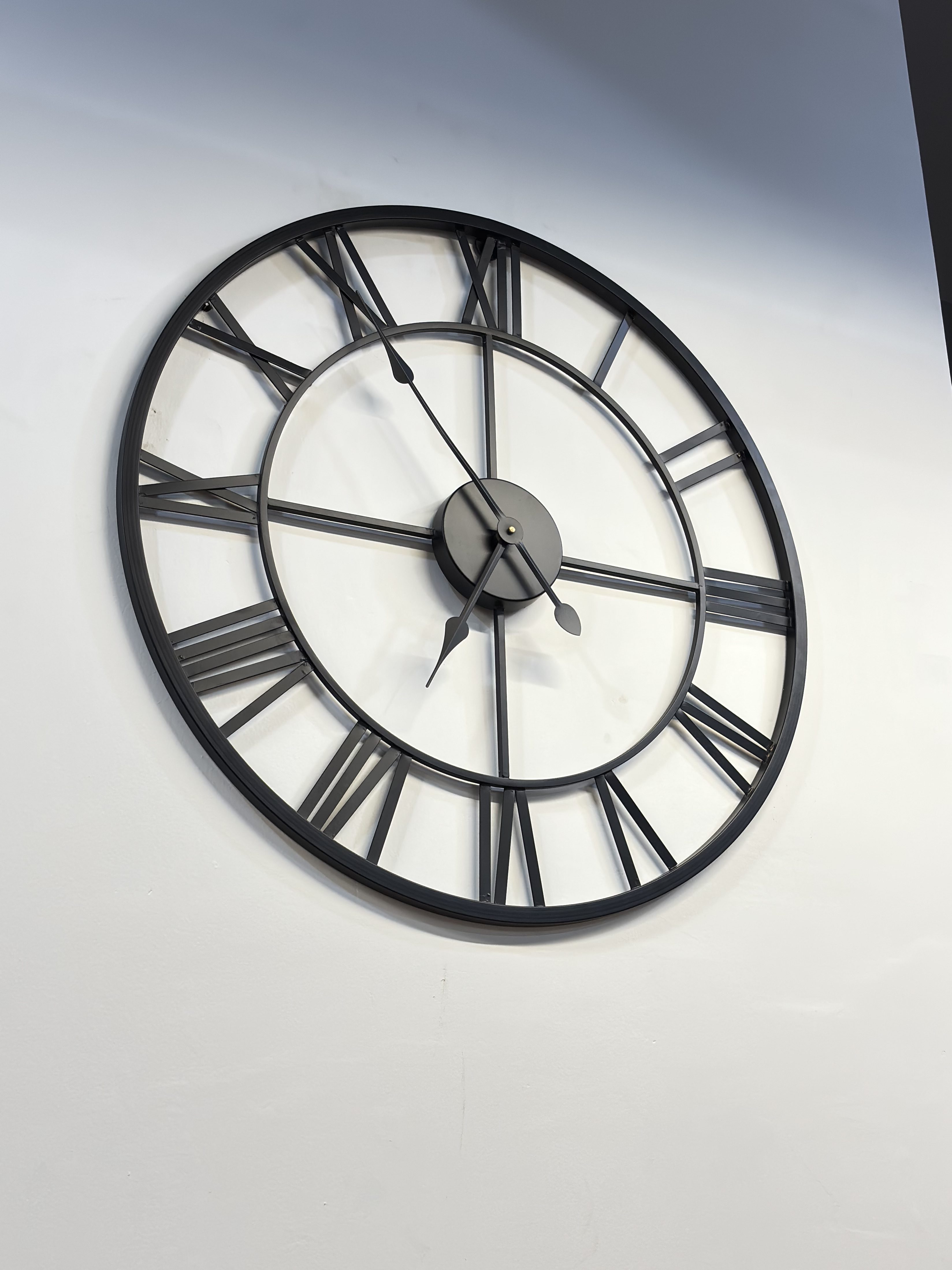 Minimalist industrial-style wall clock with a black metal frame and Roman numerals mounted on a plain white wall.