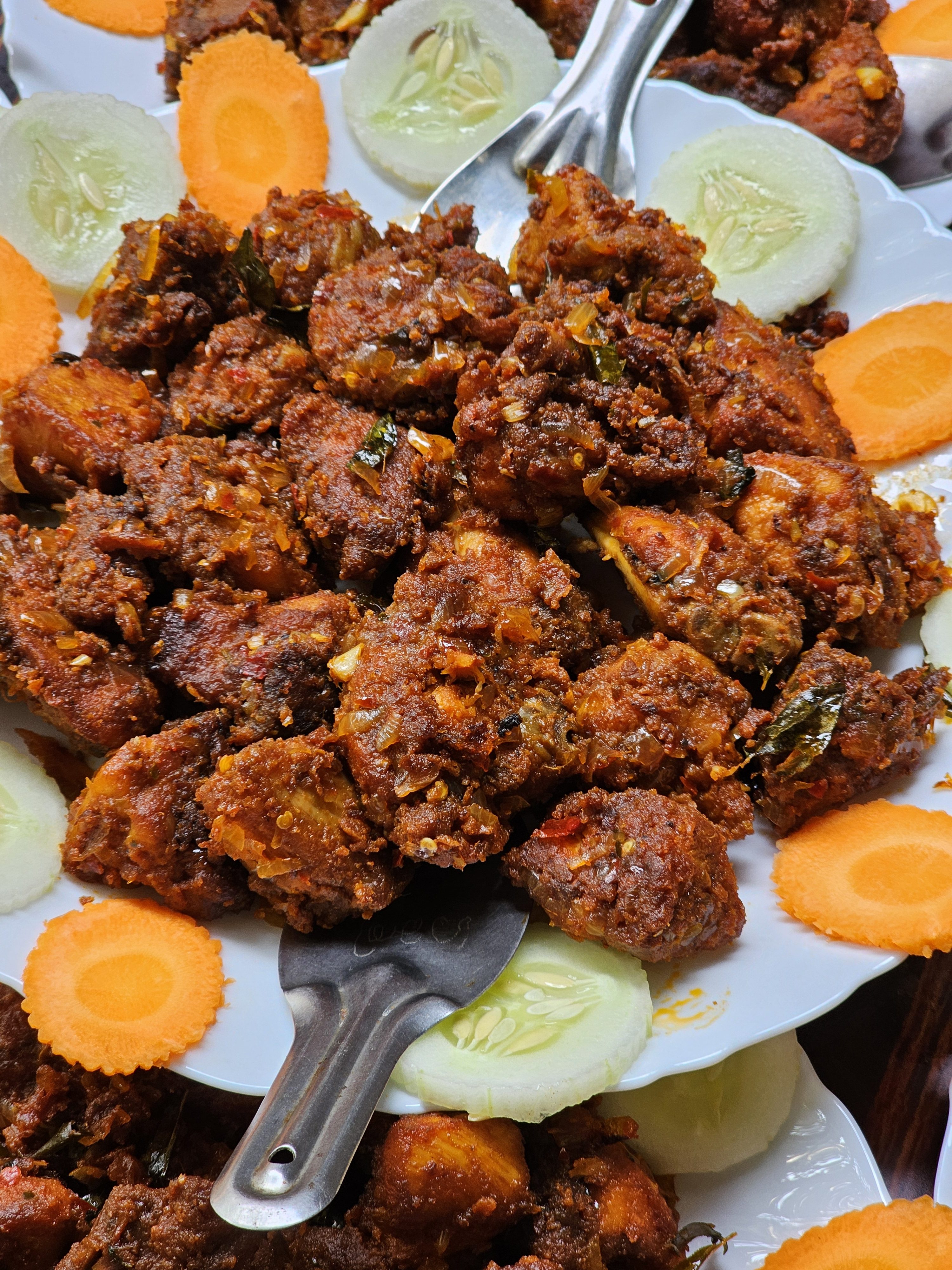 A plate of Mangalorean Chicken Sukka, aka Chicken Kondattam, a popular Indian dish, garnished with slices of cucumber and carrot. The chicken pieces are coated in a spicy red marinade and appear to be fried, and the dish is presented on a white plate with a serving spoon. Spotted in Kozhikode, Kerala.