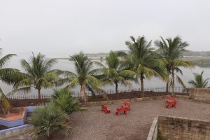 A serene outdoor setting featuring a line of coconut palm trees alongside a calm body of water. In the foreground, there are several red benches on a gravel-covered area, enclosed by stone walls and a fence. The sky appears overcast, enhancing the tranquil atmosphere.