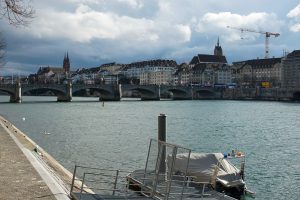 A scenic view of Rhine river with a stone bridge crossing it, surrounded by historic buildings with a church in the background. A construction crane is visible in the skyline, and swans are swimming in the river.