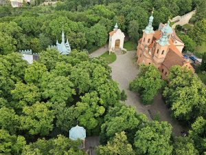 An aerial view of the Cathedral of St. Lawrence in Prague, nestled among lush greenery, within the Petřín Gardens. The cathedral, with its orange roof and towers, stands out against the surrounding trees.