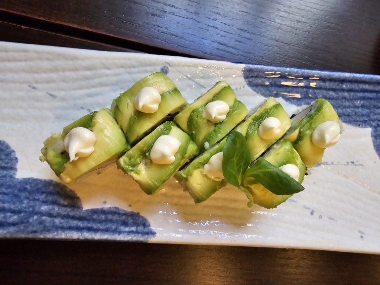 A plate of avocado sushi rolls. The sushi is arranged diagonally on a rectangular plate with a blue and white pattern. Each roll is topped with a slice of avocado and a dollop of white sauce. There is also a small green leaf on one of the sushi pieces. Captured from a restaurant in Turin, Italy.