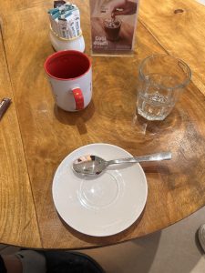 A wooden table with a red and white mug, a glass of water, a small dish with a spoon, a container of sugar packets, and a promotional card showing an iced coffee.
