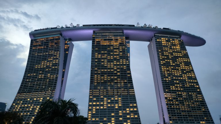 A modern high-rise building with three towers connected by a rooftop structure, illuminated at night with numerous lit windows against a cloudy sky.