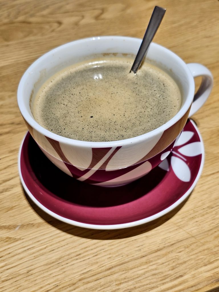 A cup of coffee with foam on top, served in a decorative white mug with maroon patterns, placed on a matching maroon saucer.