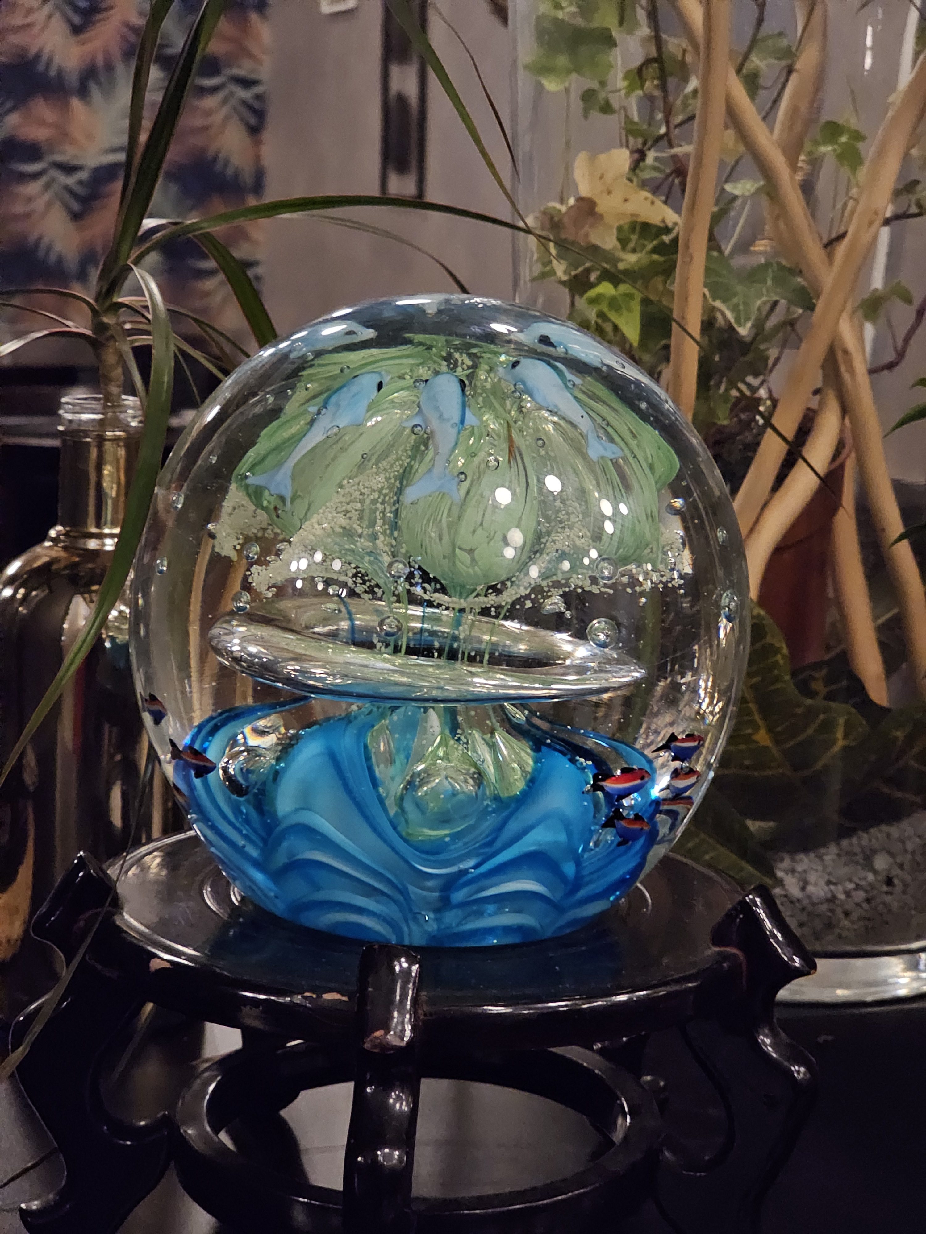 A Selkirk Glass artistic paperweight, featuring a pastel-colored magnum design. The paperweight is predominantly clear, with a green and blue design inside, resembling an abstract floral and aquatic scene. Captured from a restaurant in Turin, Italy.
