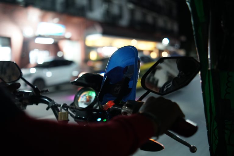 A close-up view of a person riding a motorcycle at night. The focus is on the handlebars, side mirrors, and a blue phone holder attached to the bike.