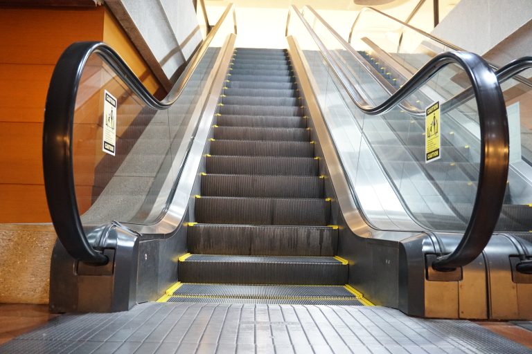 An empty escalator with black steps and yellow edges, flanked by glass panels with black handrails, leading up to another floor in a building, with caution signs on the sides.