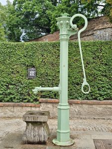 A vintage, light-green hand-operated water pump with a stone basin. The background includes a green hedge and a brick wall. From a garden in Prague. 