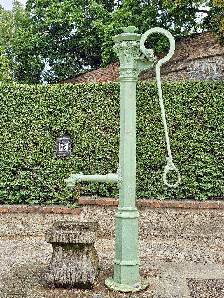 A vintage, light-green hand-operated water pump with a stone basin. The background includes a green hedge and a brick wall. From a garden in Prague.