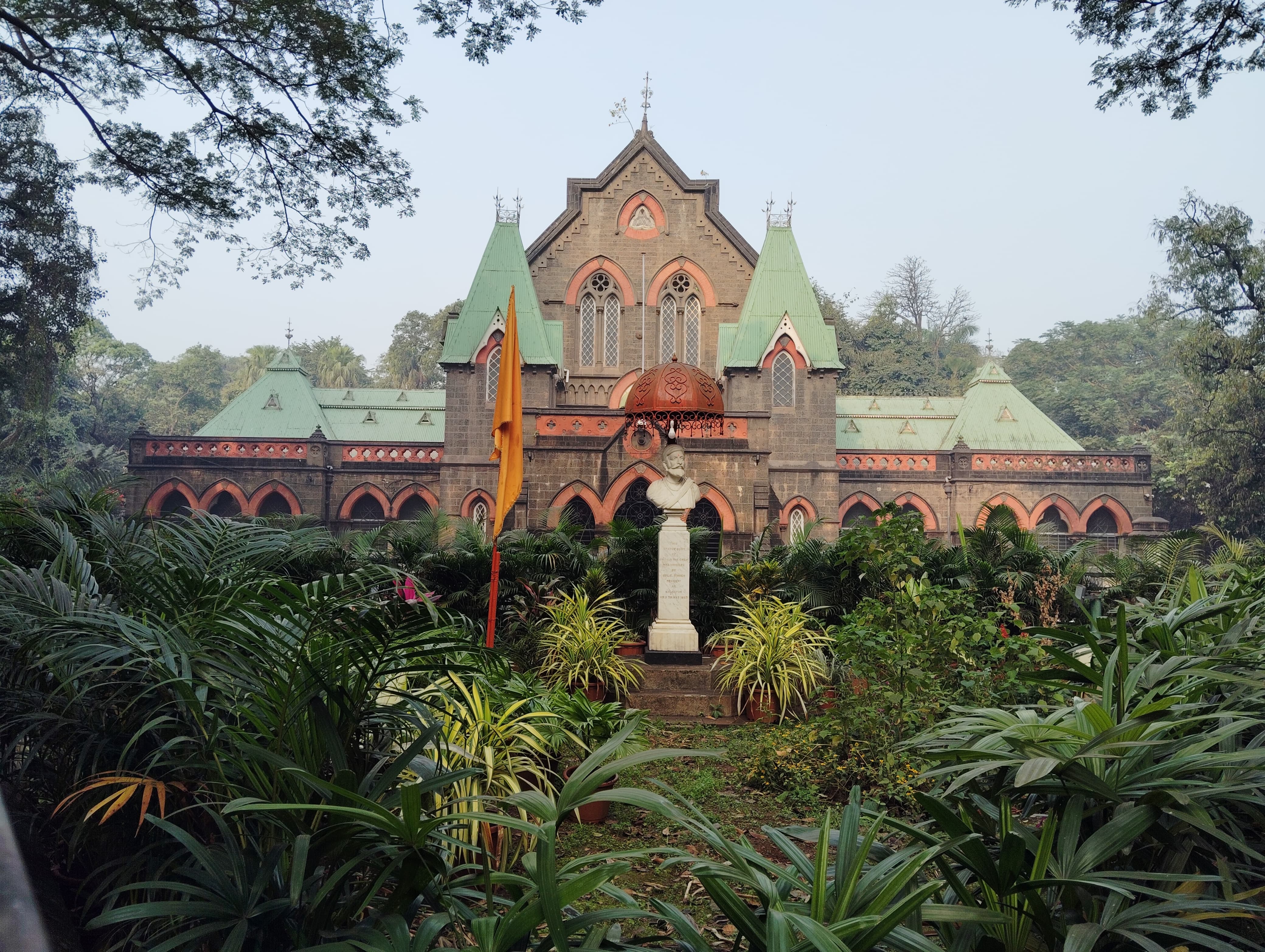 Heritage in Kolhapur - "Townhall". A historic building with gothic architecture featuring green roofs and arched windows surrounded by lush greenery. A bust on a pedestal stands in front of the building, with a flag nearby, framed by trees and foliage.
