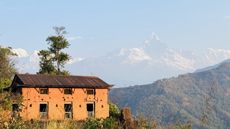 A rustic brick house with a corrugated metal roof stands amidst greenery, with a clear view of snow-capped mountains and a blue sky in the background.