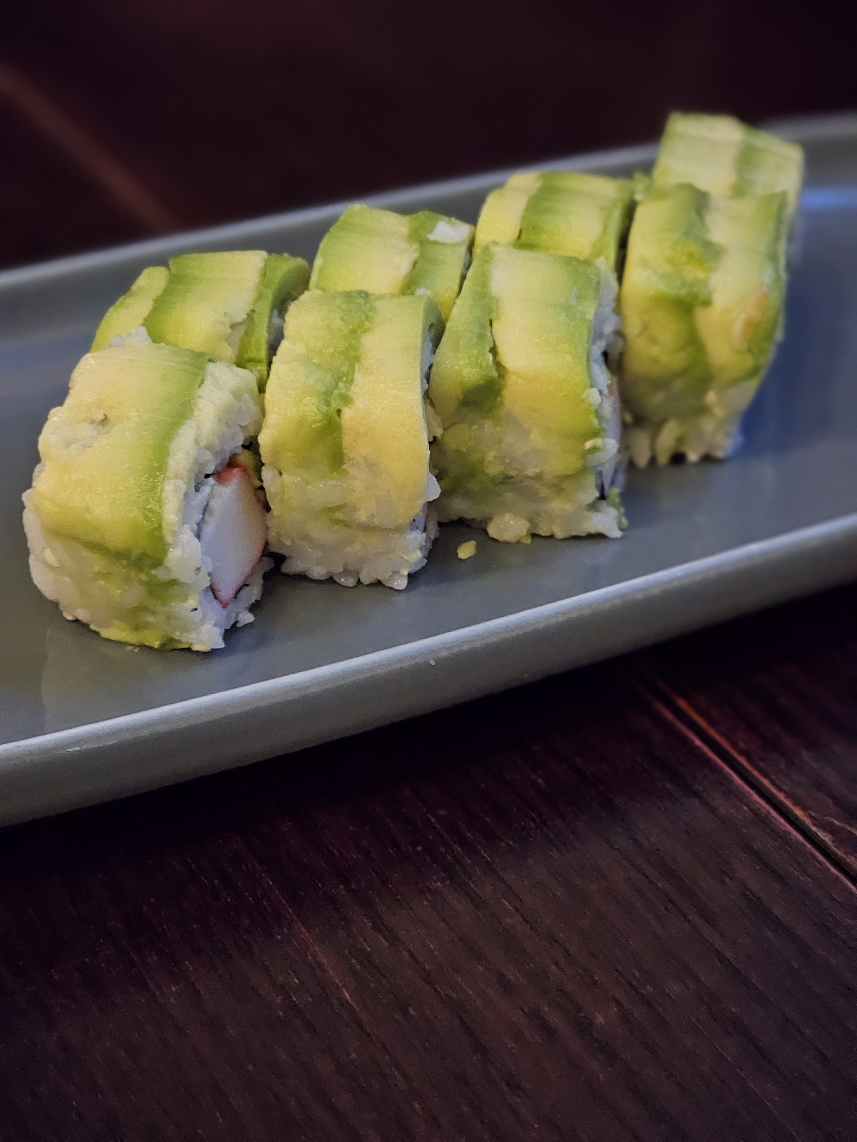 A plate of dragon roll sushi. The sushi rolls are topped with slices of avocado, crab meat, and rice. The rolls are arranged on a rectangular gray plate set on a dark wooden surface. Captured from a restaurant in Turin, Italy.