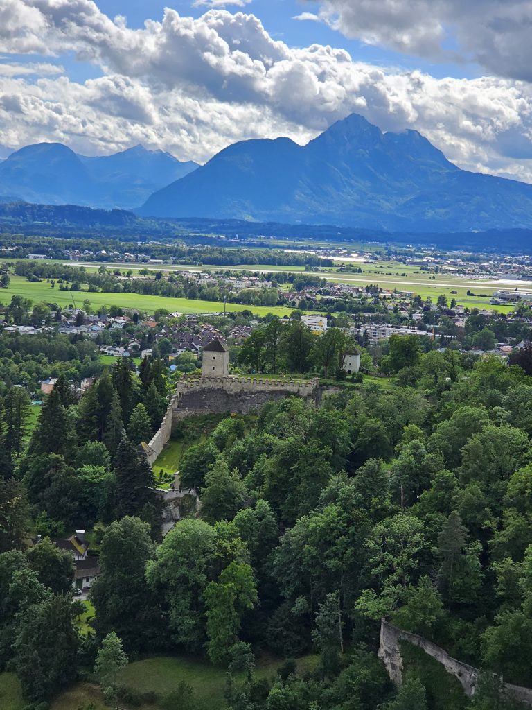 The Hohensalzburg Fortress (Salzburg, Austria) is nestled among lush greenery, with a backdrop of majestic mountains and a cloudy sky. This aerial view captures the fortress’s stone walls and towers, highlighting its historical architecture and scenic surroundings.