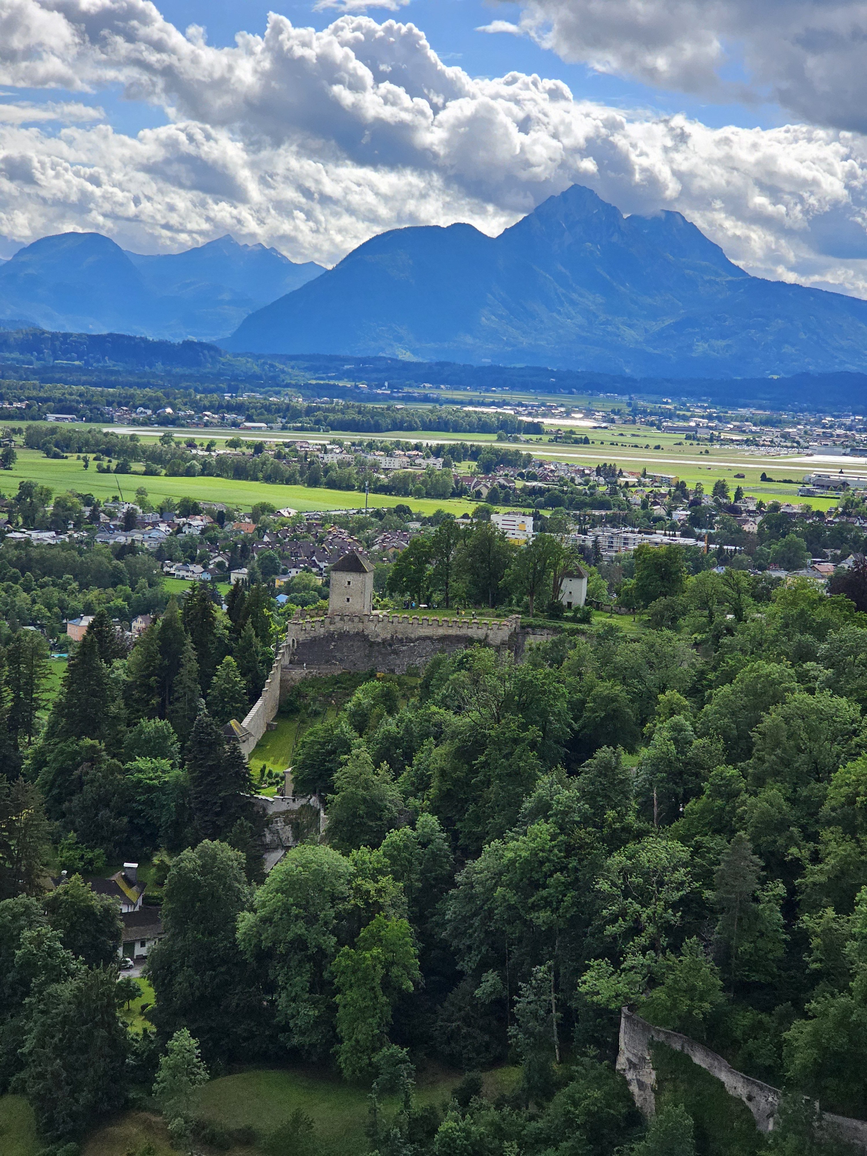 The Hohensalzburg Fortress (Salzburg, Austria) is nestled among lush greenery, with a backdrop of majestic mountains and a cloudy sky. This aerial view captures the fortress's stone walls and towers, highlighting its historical architecture and scenic surroundings.
