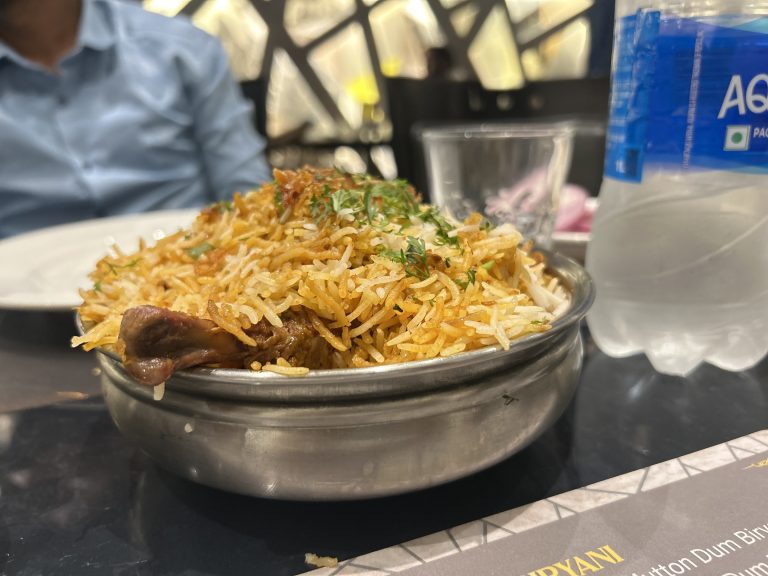 A bowl of flavorful biryani garnished with herbs, served on a table alongside a clear glass and a bottle of water. A person in a blue shirt is partially visible in the background.